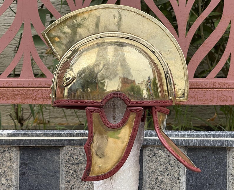 K&ouml;nnte beinhalten: Ein polierter r&ouml;mischer Helm aus Messing mit gew&ouml;lbter Oberseite und roten Lederakzenten. Der Helm hat Wangenklappen und einen dekorativen Kamm, der historische Handwerkskunst zeigt. Der Helm ist eine Nachbildung antiker Milit&auml;rhelme.