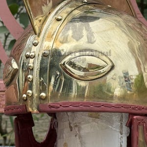 K&ouml;nnte beinhalten: Nahaufnahme eines Messinghelms mit poliertem Finish. Der Helm hat augenf&ouml;rmige Aussparungen und ist mit Nieten und einer roten Lederausstattung versehen. Das Design deutet auf ein historisches oder Fantasy-Thema hin.