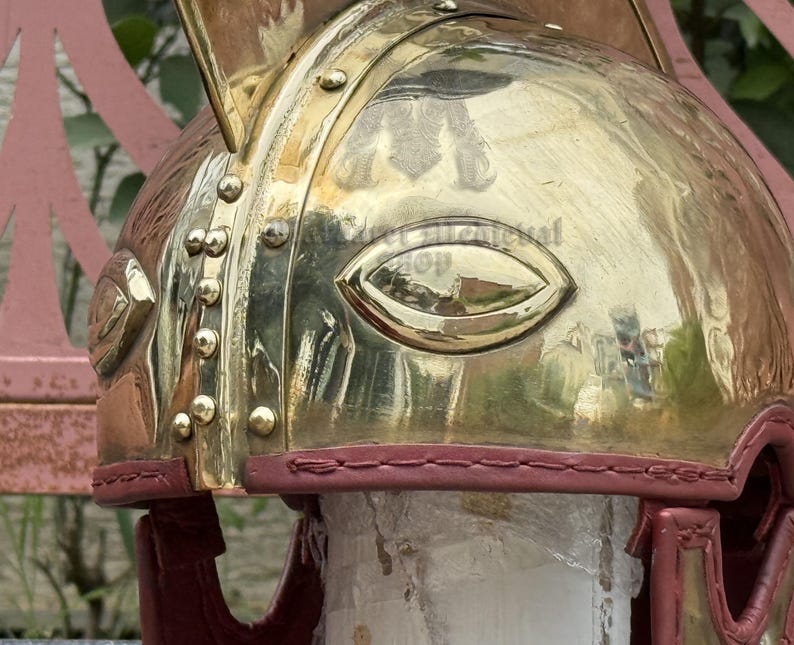 K&ouml;nnte beinhalten: Nahaufnahme eines Messinghelms mit poliertem Finish. Der Helm hat augenf&ouml;rmige Aussparungen und ist mit Nieten und einer roten Lederausstattung versehen. Das Design deutet auf ein historisches oder Fantasy-Thema hin.