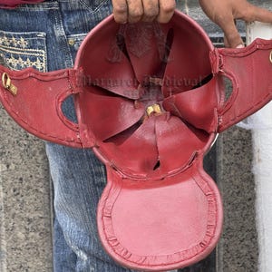 K&ouml;nnte beinhalten: Ein roter Lederhelm mit fl&uuml;gelartigen Seitenklappen und goldfarbenen Metallakzenten. Das Innere des Helms ist sichtbar und zeigt die N&auml;hte und Konstruktionsdetails. Der Helm wird von einer Person gehalten.
