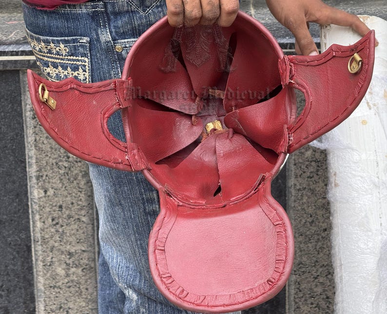 K&ouml;nnte beinhalten: Ein roter Lederhelm mit fl&uuml;gelartigen Seitenklappen und goldfarbenen Metallakzenten. Das Innere des Helms ist sichtbar und zeigt die N&auml;hte und Konstruktionsdetails. Der Helm wird von einer Person gehalten.