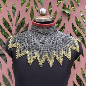 Könnte beinhalten: Ein Kettenkragen mit hohem Kragen, ausgestellt auf einer schwarzen Schaufensterpuppe. Der Kragen ist hauptsächlich silberfarben, mit einem dekorativen goldfarbenen Rand. Am Halsansatz ist ein rotes Lederband zu sehen.