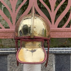 K&ouml;nnte beinhalten: Ein polierter Messinghelm mit einem markanten zentralen Dorn. Der Helm hat eine abgerundete Krone und einen Gesichtsschutz mit roter Lederausstattung. Das Wort "Medieval" ist auf dem Gesichtsschutz zu sehen.