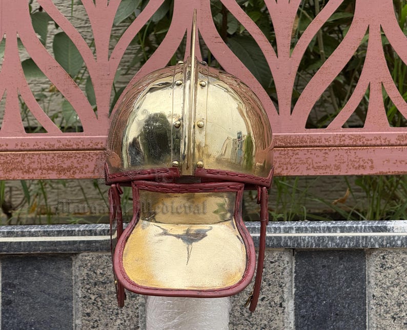 K&ouml;nnte beinhalten: Ein polierter Messinghelm mit einem markanten zentralen Dorn. Der Helm hat eine abgerundete Krone und einen Gesichtsschutz mit roter Lederausstattung. Das Wort "Medieval" ist auf dem Gesichtsschutz zu sehen.