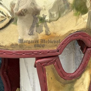 K&ouml;nnte beinhalten: Nahaufnahme eines dekorativen mittelalterlichen Helms mit goldenen und roten Akzenten. Der Helm weist ein detailliertes Design und den Text "Margaret Medieval Shop" auf. Der Helm ist wahrscheinlich ein Sammler- oder Dekorationsartikel.