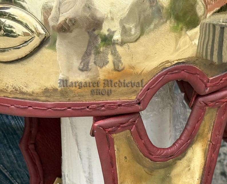 K&ouml;nnte beinhalten: Nahaufnahme eines dekorativen mittelalterlichen Helms mit goldenen und roten Akzenten. Der Helm weist ein detailliertes Design und den Text "Margaret Medieval Shop" auf. Der Helm ist wahrscheinlich ein Sammler- oder Dekorationsartikel.