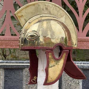 K&ouml;nnte beinhalten: Ein r&ouml;mischer Helm aus Messing mit poliertem Finish. Der Helm hat einen gebogenen Kamm, Augen&ouml;ffnungen und Wangenklappen. Die Wangenklappen sind tiefrot und der Helm ist auf einem wei&szlig;en zylindrischen Objekt montiert.