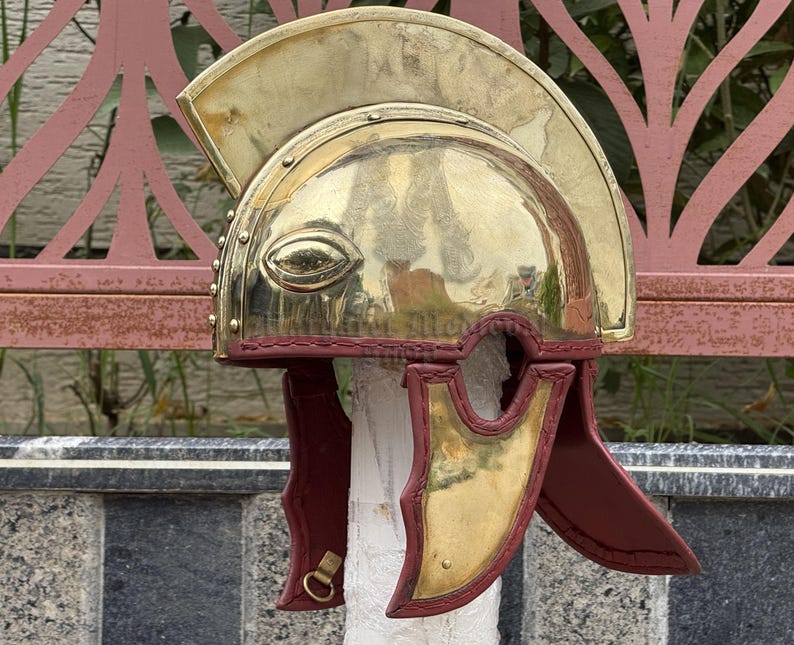 K&ouml;nnte beinhalten: Ein r&ouml;mischer Helm aus Messing mit poliertem Finish. Der Helm hat einen gebogenen Kamm, Augen&ouml;ffnungen und Wangenklappen. Die Wangenklappen sind tiefrot und der Helm ist auf einem wei&szlig;en zylindrischen Objekt montiert.