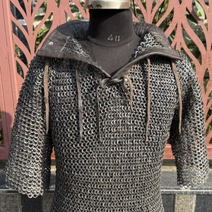 May include: A chainmail shirt with a leather collar and short sleeves. The metal rings are interlocked, creating a flexible, protective garment. The collar is dark brown, and the shirt is displayed on a black mannequin.