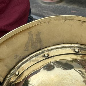 K&ouml;nnte beinhalten: Nahaufnahme eines goldfarbenen Metallhelms mit gebogenem Rand. Der Helm hat eine reflektierende Oberfl&auml;che und ist mit Nieten und einem gepr&auml;gten Emblem verziert. Der Text "Mara Metal Shop" ist auf dem Helm sichtbar.