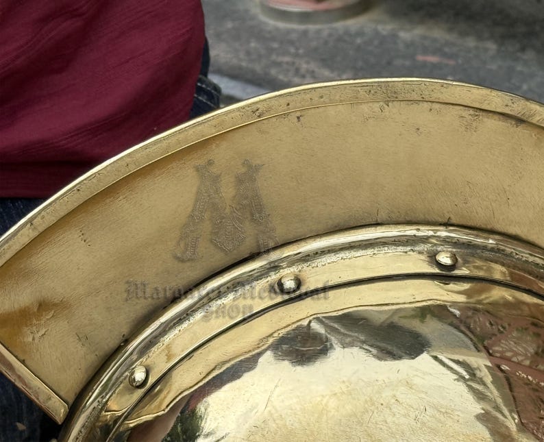 K&ouml;nnte beinhalten: Nahaufnahme eines goldfarbenen Metallhelms mit gebogenem Rand. Der Helm hat eine reflektierende Oberfl&auml;che und ist mit Nieten und einem gepr&auml;gten Emblem verziert. Der Text "Mara Metal Shop" ist auf dem Helm sichtbar.