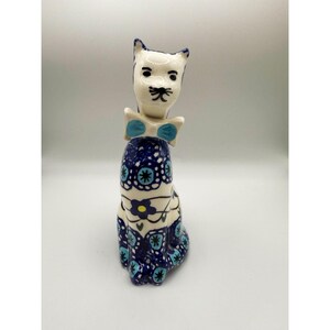 May include: A ceramic cat figurine featuring a white base with blue and dark blue floral patterns. The cat has a light blue bow tie around its neck, painted black eyes, a nose, and whiskers. The body is decorated with repeating circular motifs.