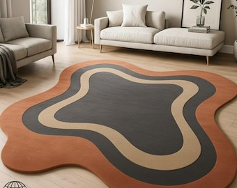Puede incluir: Una alfombra moderna y abstracta con una forma orgánica única. La alfombra presenta un centro gris oscuro, bordeado por una onda beige y una capa exterior de color naranja quemado. La alfombra está colocada sobre un suelo de madera clara.