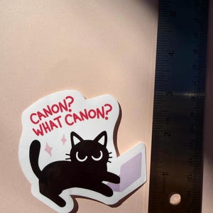 May include: A sticker featuring a black cat with the text "CANON? WHAT CANON?" in red. The cat is drawn with a laptop. The sticker is approximately 3 inches in length.