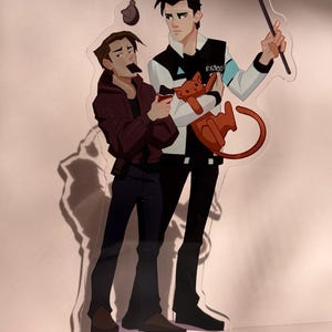 Könnte beinhalten: Ein klarer Acrylständer mit zwei illustrierten Figuren. Eine Figur hält ein Katzenplüschtier, die andere einen kleinen Gegenstand. Die Figur rechts trägt eine schwarz-weiße Jacke mit dem Aufdruck "RK900". Über den Figuren befinden sich eine Angelrute und eine Pose.