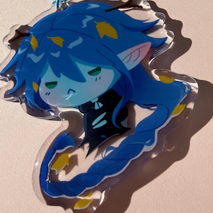 May include: A clear acrylic charm depicting a blue anime-style character with long blue hair and yellow details. The charm has a blue clasp, possibly for use as a keychain or accessory.