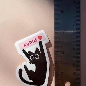 May include: A black cat sticker with the word "KUDOS" in red on a pink banner, and a red heart. The sticker is approximately 3 inches tall. The cat has large, round eyes and is giving a thumbs-up gesture.