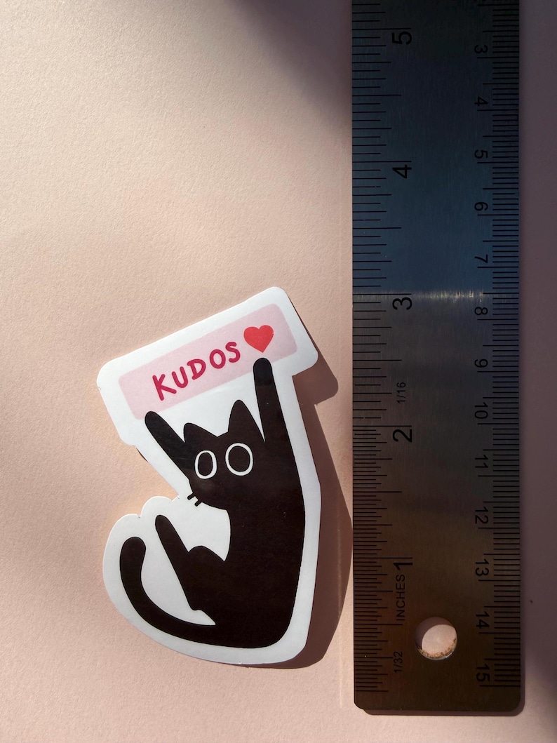 May include: A black cat sticker with the word "KUDOS" in red on a pink banner, and a red heart. The sticker is approximately 3 inches tall. The cat has large, round eyes and is giving a thumbs-up gesture.