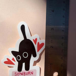 May include: A sticker featuring a black cat with white eyes and red heart accents. The cat is arched with its tail up. The sticker is approximately 2 inches tall and has the word "SLOWBURN" in pink on a white rectangle.