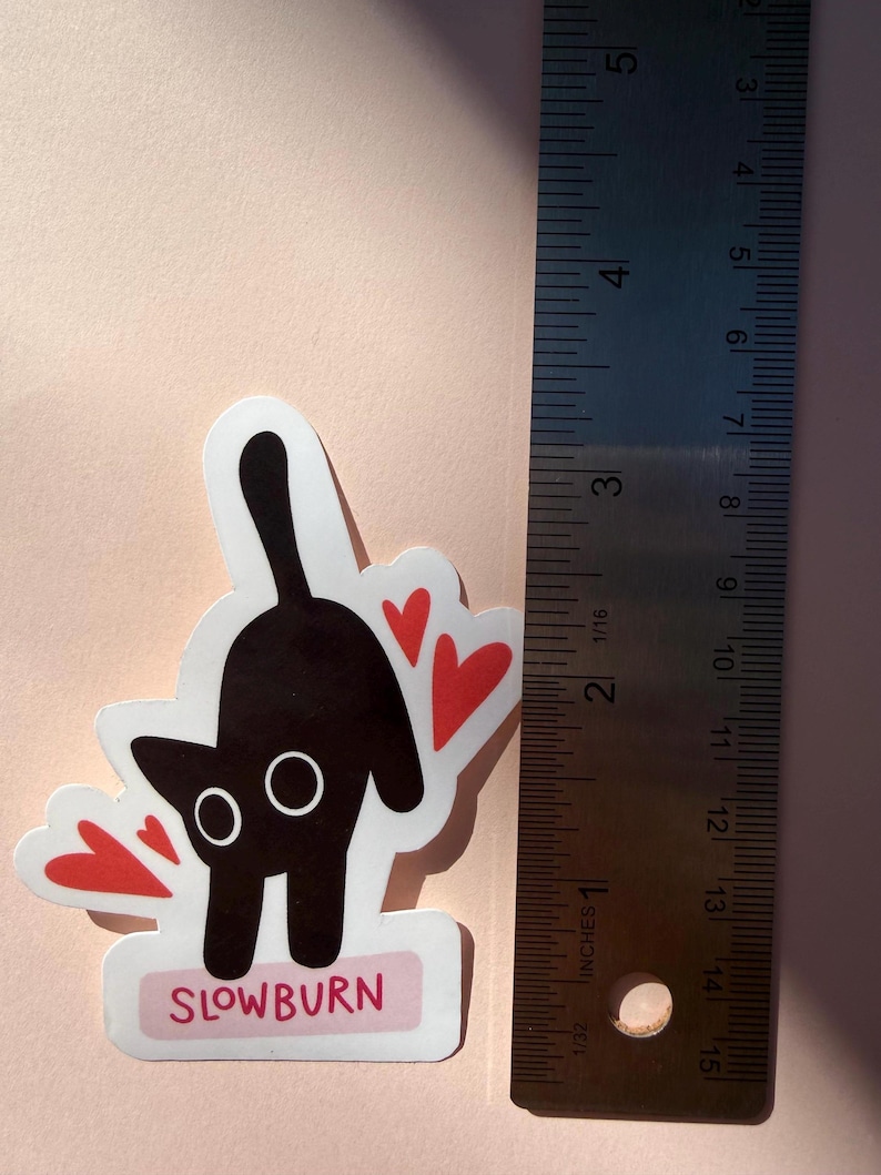 May include: A sticker featuring a black cat with white eyes and red heart accents. The cat is arched with its tail up. The sticker is approximately 2 inches tall and has the word "SLOWBURN" in pink on a white rectangle.