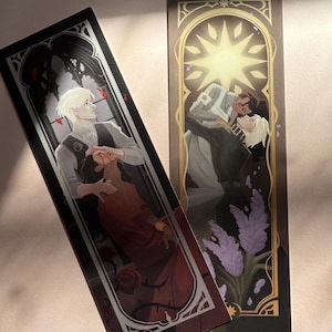 Puede incluir: Dos marcapáginas rectangulares con ilustraciones. Uno presenta a un hombre con una túnica roja, el otro representa a dos hombres abrazándose bajo un sol dorado. Ambos tienen bordes ornamentados y están sobre un fondo claro.