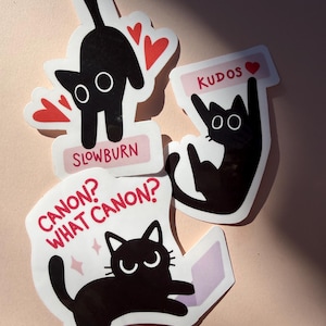 May include: Four cat stickers with black cat illustrations. One sticker has the text "SLOWBURN" and red hearts. Another says "KUDOS" with a heart. The third sticker says "CANON? WHAT CANON?" with a cat and a book.