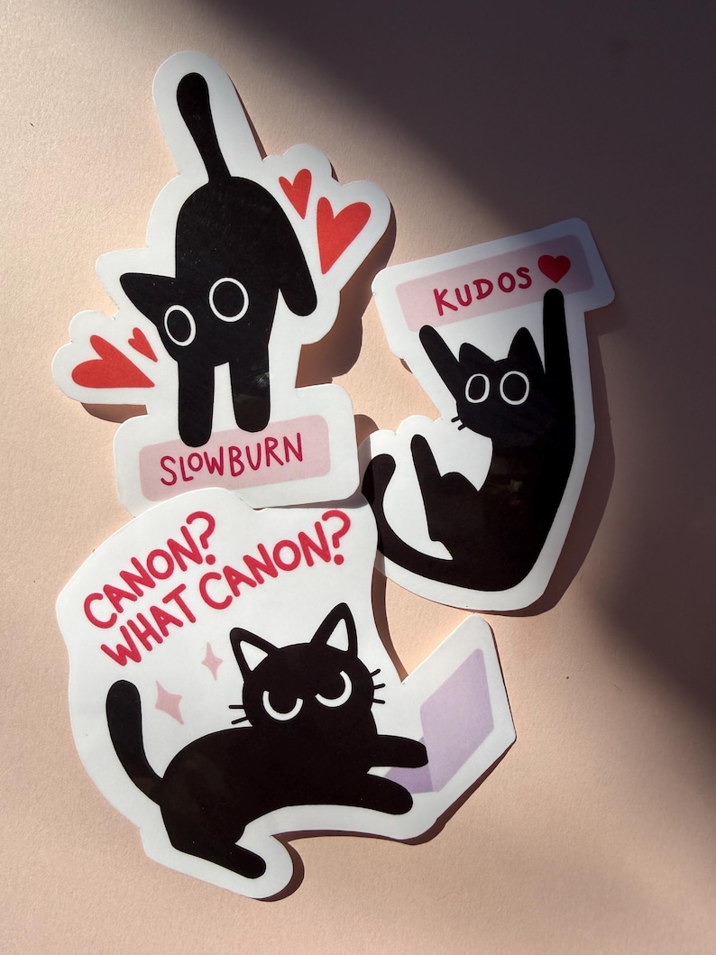 May include: Four cat stickers with black cat illustrations. One sticker has the text "SLOWBURN" and red hearts. Another says "KUDOS" with a heart. The third sticker says "CANON? WHAT CANON?" with a cat and a book.