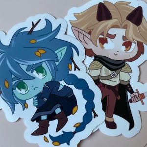 Könnte beinhalten: Zwei Aufkleber mit Comicfiguren. Einer ist ein blauhäutiger Elf mit grünen Augen und einem blauen Zopf. Der andere ist ein blonder Elf mit roten Augen und braunen Hörnern. Beide tragen Kleidung und halten Waffen.