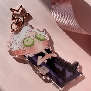 Könnte beinhalten: Ein durchsichtiger Acryl-Schlüsselanhänger mit einer Comicfigur mit weißen Haaren, grünen Gurkenscheiben über den Augen und einem lila-schwarzen Outfit. Die Figur sitzt auf einem Stuhl mit einer kleinen Box davor.