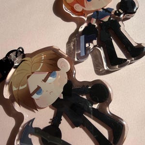 May include: Two clear acrylic charms with cartoon illustrations of a character with blonde hair, blue eyes, and black clothing. One charm holds a knife and a gun, while the other holds a hatchet and a black object. Both charms have a black keychain attachment.