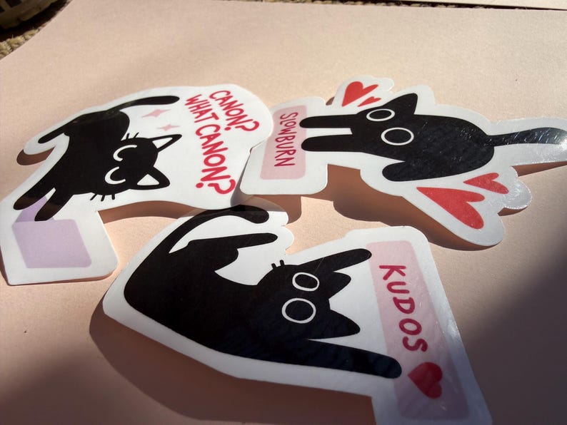 May include: Three die-cut stickers featuring black cat illustrations. One sticker reads "Canon? What Canon?" in red text. Another says "Slowburn" and the third says "Kudos". Each sticker has a white background and a pink border.
