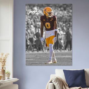 May include: A framed print of a football player in a maroon and gold uniform with the number 0. The player is wearing a helmet and standing on a field. The background is in black and white.