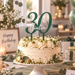Op de afbeelding: Een donkergroene taarttopper met het getal 30 en de naam Giulia in witte schrijfletters in het midden. De topper staat op een ronde taart met wit glazuur, versierd met witte bloemen, groene blaadjes en gehakte pistachenootjes.