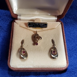 May include: A vintage jewellery set presented in a velvet-lined box. The set features a gold-toned necklace with a heart pendant and matching earrings, each with a genuine ruby. The box is marked "JCPenney" and the necklace tag reads "Genuine Ruby 14kt. Gold Filled."