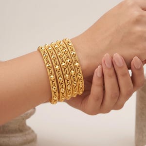 May include: A set of five gold bangles featuring a repeating geometric pattern with small, raised, polished circular accents along the centre. The bracelets have a textured, beaded edge design and a bright, reflective metallic finish.