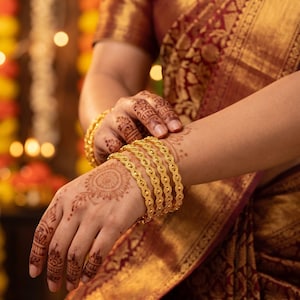 May include: A set of four gold bangles featuring a repeating oval and dot pattern. The bangles are worn on a wrist adorned with intricate henna designs, paired with a deep maroon and gold fabric garment that has a detailed woven border.