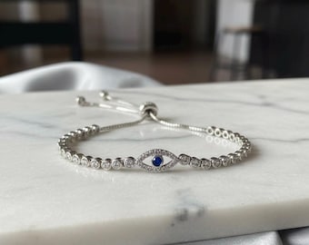 Pode incluir: Pulseira de prata com um design central de olho grego. A pulseira apresenta uma fileira de pequenas pedras transparentes ao redor do pulso e uma pedra azul no centro do olho. A pulseira é exibida em uma superfície de mármore branco.