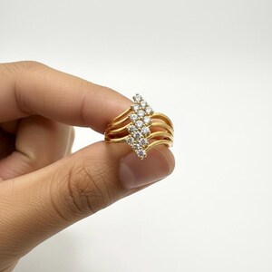 Puede incluir: Un anillo dorado con un grupo de pequeñas piedras preciosas transparentes. El anillo tiene un diseño multibanda con una disposición central de piedras que apunta hacia arriba. El anillo se sostiene sobre un fondo blanco.