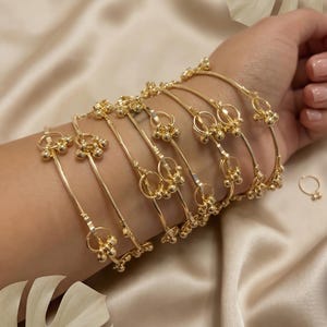 Puede incluir: Se muestran pulseras doradas y un anillo en una muñeca. Las pulseras tienen un diseño detallado con pequeñas decoraciones esféricas, y el anillo tiene un estilo a juego. Las joyas se presentan sobre un fondo de tela sedosa de color crema.