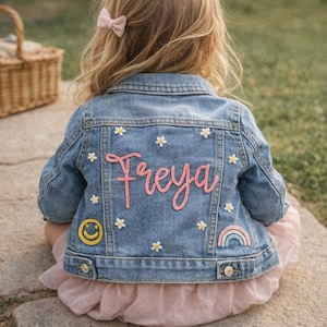 May include: A light blue denim jacket featuring the name Freya embroidered in pink script on the back. The jacket is decorated with small white flower embroidery, a yellow smiley face patch, and a small rainbow patch on the lower back panel.