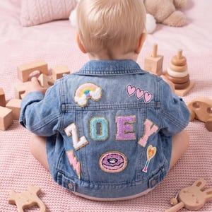 May include: A light blue denim jacket featuring colourful patches on the back. The patches include a rainbow, three pink hearts, a lightning bolt, a doughnut, a star wand, and the name ZOEY spelled out in large, textured, multi-coloured varsity letters.