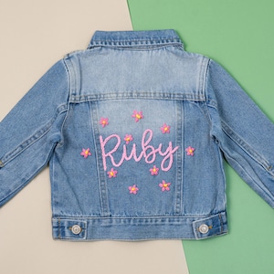 May include: A light blue denim jacket featuring the name "Ruby" embroidered in pink script on the back, with pink flower designs. The jacket has a classic collar, button fastenings, and long sleeves.