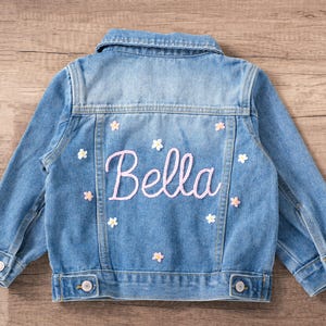 May include: Pale blue denim jacket with the name "Bella" embroidered in pink script on the back. The jacket is decorated with small white and pink flower appliqués. Features button closures and a classic collar.