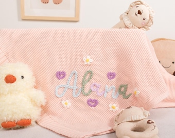May include: A pale pink baby blanket embroidered with the name "Alana" in pastel shades, decorated with small flower and heart motifs. A plush chick, lion, and other toys are arranged around the blanket.