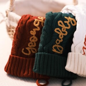 Puede incluir: Tres gorros de punto trenzado en tonos rojo, verde y blanco, cada uno con un nombre bordado en un color contrastante. El gorro rojo dice "George", el gorro verde dice "Daddy", y el gorro blanco tiene un nombre bordado en rojo. Una cesta de mimbre está en el fondo.