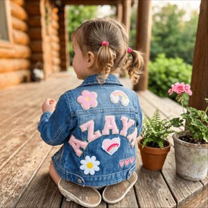 Könnte beinhalten: Eine blaue Jeansjacke mit dem Namen AZAYA in großen rosa Buchstaben auf dem Rücken. Die Jacke ist mit mehreren Aufnähern verziert, darunter eine rosa Blume, ein Regenbogen, ein weißes Gänseblümchen, ein Herz mit Regenbogen und drei kleine rosa Herzen.