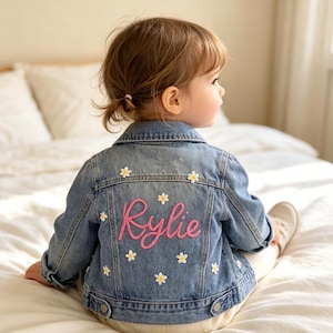 May include: A blue denim jacket featuring the name Rylie embroidered in pink script on the back, surrounded by small white and yellow daisy flower appliques. The jacket has a classic collar, long sleeves, and buttoned cuffs and hem details.