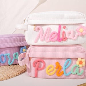 以下が含まれることがあります： 白、ピンク、ラベンダー色のコーデュロイコスメティックバッグ3つ。各バッグには、カラフルな盛り上がった糸文字で名前と糸の花が特徴です。白いバッグはピンクで「Melia」、ピンクのバッグは虹色で「Perla」、ラベンダーのバッグは青と緑で「Flora」と書かれています。