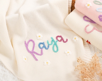May include: Cream knitted baby blanket embroidered with the name "Raya" in colourful thread. The blanket features small white daisy flowers. Other folded blankets with floral embroidery are also visible.