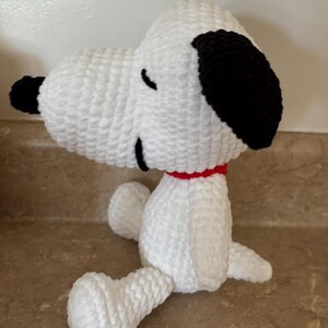 May include: A handmade crochet Snoopy plush toy. The plush is white with black ears and a black nose. It has a red collar and is sitting down. The toy is made of soft yarn and is a popular character from the Peanuts comic strip.